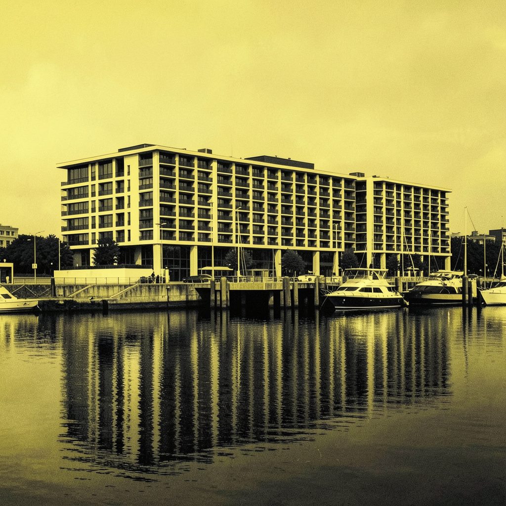 Hard two-tone duotone photograph of a modern waterfront hotel facade reflected on still harbor water in muted gold and deep blue tones